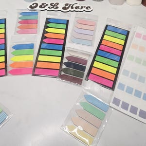May include: A collection of adhesive page markers and index tabs in various shapes and colors, including neon arrow-shaped strips, pastel rectangular labels, and pencil-patterned flags, all packaged in clear plastic sleeves with the text FANDAN visible.