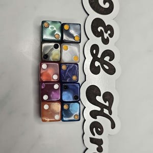 May include: Nine multi-colored dice with white pips and gold or black dots. The dice are arranged in a 3x3 grid. A white and brown sign with the words "Here & Now" is on the right side.