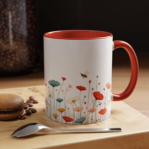 May include: A white ceramic mug with a red rim and handle. The mug features a colorful floral design with a bee flying above the flowers.