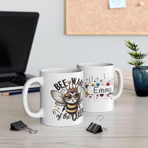 May include: White ceramic mug with a bee wearing a crown and sunglasses. The mug says "Bee-ware of the Queen" and has a floral design with the name "Emma".