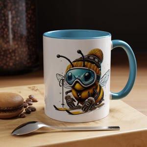 Puede incluir: Taza de cerámica blanca con borde y asa azules. La taza presenta una ilustración de dibujos animados de una abeja con un gorro de invierno y gafas de esquí, esquiando cuesta abajo por una montaña nevada.