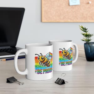 May include: Two white ceramic mugs with a rainbow and bee design. The mugs have the text "Be Proud" on them.