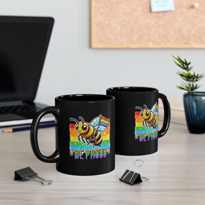May include: Two black ceramic mugs with a rainbow pride flag and a cartoon bee design. The text "Be Proud" is printed on the mugs.