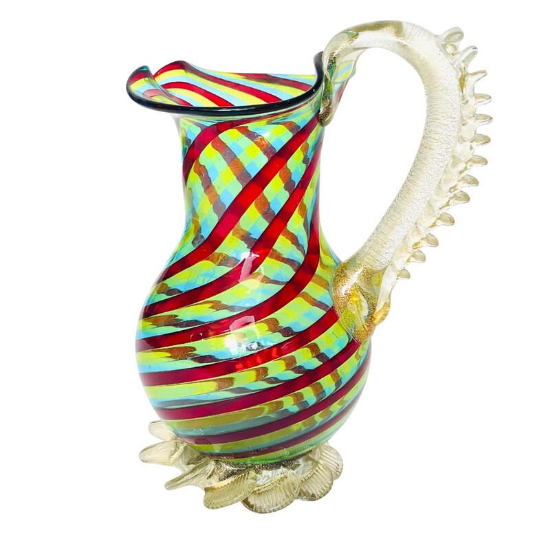 May include: A colorful glass pitcher with a decorative handle and base. The pitcher features a vibrant pattern of red, yellow, and blue stripes. The handle is clear with gold accents, and the base has a flower-like design.