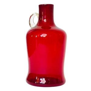 Może przedstawiać: Żywy, czerwony szklany dzbanek z przezroczystym uchwytem. Dzbanek ma zaokrąglony korpus i wąską szyjkę. Uchwyt jest przezroczysty i przymocowany u góry, zakrzywiając się w dół, aby połączyć się z korpusem. Szkło jest gładkie i odbija światło.