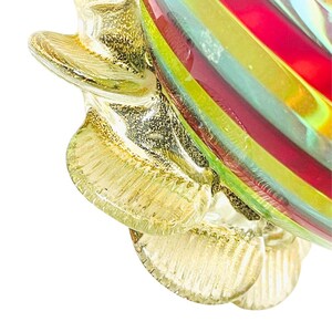 May include: Close-up of a decorative glass object with a petal-like design. The petals are a translucent gold color with glitter accents. The interior of the object features bands of red, yellow, green, and blue glass.