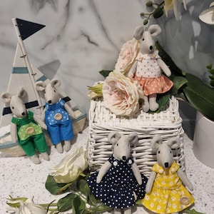 May include: A group of fabric mouse dolls, each wearing a different outfit. The dolls are positioned near a small wooden sailboat and a white wicker basket filled with flowers. The mice are dressed in dresses and dungarees in various colours and patterns.