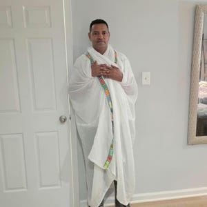 May include: A white traditional Ethiopian netela shawl made of lightweight fabric, draped over the shoulders and chest, featuring a decorative border with colorful geometric patterns in red, green, yellow, and blue along the edges of the garment.