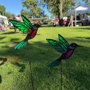 May include: Two metal hummingbird garden stakes with green and black wings, red and purple accents, and long, thin metal rods for placement in the ground. The stakes are in a grassy area with people and tents in the background.
