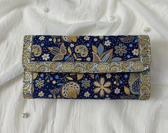 May include: A rectangular wallet with a navy blue background and a floral pattern in gold, white, and light blue. The wallet has a fold-over flap and a gold border. The design includes flowers, leaves, and paisley-like shapes.