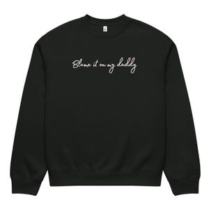 Puede incluir: Sudadera negra de cuello redondo con la frase "Blame it on my daddy" en escritura cursiva blanca en el pecho. La sudadera tiene dobladillo y puños acanalados.