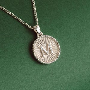 Könnte beinhalten: Eine silberne Halskette mit einem runden Anhänger, der den Buchstaben "M" in erhabener Form zeigt. Der Anhänger hat ein strukturiertes, sonnenstrahlenartiges Muster um den Buchstaben herum.