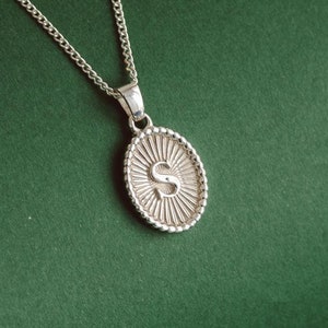 Op de afbeelding: Een zilveren ketting met een zilveren ovale hanger met de letter "S" in reliëf.