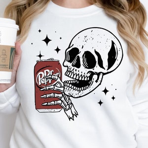 May include: White sweatshirt with a graphic of a skull drinking a can of Dr Pepper. The skull is black and white with a red can. There are small white stars around the skull.