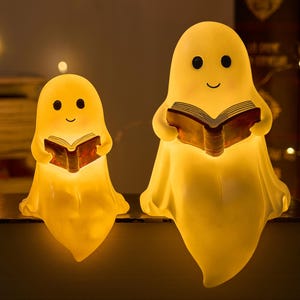 May include: Two illuminated ghost-shaped lamps. Each ghost figure holds a book and has a smiling face. The lamps are yellow and emit a warm glow, creating a cosy atmosphere. Ideal for Halloween or a whimsical touch.