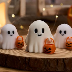 May include: Three white ghost figurines with black eyes and smiles, each holding an orange jack-o'-lantern bucket. The ghosts are on a wooden slice, with a blurred background of candles and lights, creating a Halloween theme.