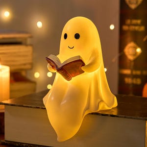 Op de afbeelding: Een gloeiende, gele nachtlamp in de vorm van een spook dat een boek met een bruine kaft leest. De lamp heeft een lachend gezicht en zit op een groot boek. Een brandende kaars en andere boeken staan op de achtergrond.