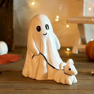 Op de afbeelding: Een witte keramische spookfiguur met zwarte ogen en een lachende mond, die een riem vasthoudt die is bevestigd aan een kleine witte hondenfiguur. De figuren staan op een houten oppervlak, met pompoenen en decoratieve lampjes op de achtergrond.