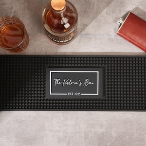 May include: A black bar mat with a textured surface, featuring a white framed rectangle with the text "The Kelvin's Bar EST. 2025". A glass of amber liquid, a liquor bottle, and a red flask are also visible.
