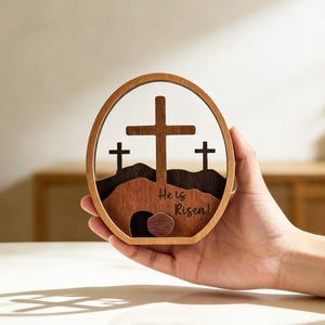 May include: Wooden Easter decoration featuring three crosses on a hill, with the text "He is Risen!" The design is set within an oval frame. The piece is made of layered wood in shades of brown and tan.