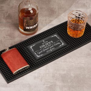 May include: A black rubber bar mat with a personalized sign that reads "COLTEN'S BEST BAR Good Friend Good Drinks GREAT TIMES". A red leather flask, a glass of amber liquid, and a whiskey bottle are on the mat.