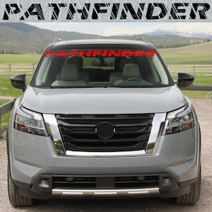 Puede incluir: Un SUV gris con una pancarta roja "PATHFINDER" en el parabrisas. El vehículo tiene una parrilla negra, detalles cromados y un círculo negro en el centro. El fondo muestra una cordillera y una valla de madera.