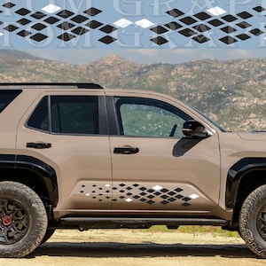 Op de afbeelding: Een beige SUV met zwarte accenten en ruitvormige stickers. Het voertuig heeft zwarte wielen met rode wieldoppen en staat op een onverharde weg met een berglandschap. De tekst "CUSTOM GRAPHICS" staat bovenaan.