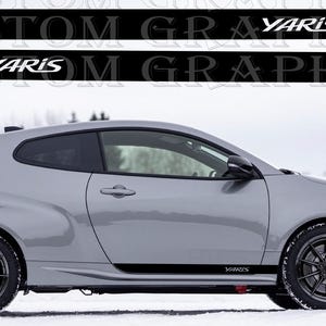Op de afbeelding: Een zilveren Toyota Yaris GR sportwagen met zwarte accenten en wielen staat geparkeerd in de sneeuw. Zwarte stickers met het woord "Yaris" staan op de zijkant. De auto heeft rode remklauwen.
