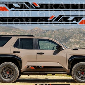 Peut inclure: Un SUV beige avec des détails noirs et des pneus tout-terrain. Le véhicule présente des décalcomanies graphiques noires et rouges sur le côté. L'arrière-plan montre une chaîne de montagnes et le texte "CUSTOM GRAPHICS".