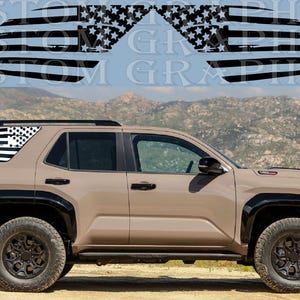 Könnte beinhalten: Ein beigefarbener SUV mit schwarzen Akzenten und Geländereifen. Das Heckfenster zeigt eine schwarz-weiße amerikanische Flaggen-Grafik. Über dem Fahrzeug sind die Worte "CUSTOM GRAPHICS" zu sehen.