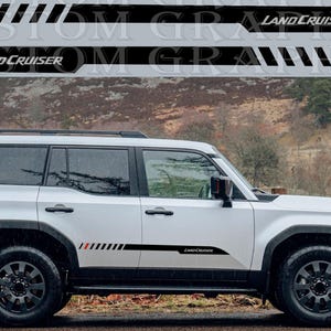 May include: White SUV with black accents and Land Cruiser decals. The decals are black with red and white stripes. The vehicle has black wheels and a roof rack. The background is a blurred outdoor scene.