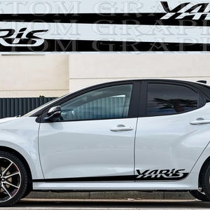 Puede incluir: Toyota Yaris blanco con detalles en negro y llantas de aleación negras y rojas. El coche tiene franjas laterales negras con la palabra "YARIS" en blanco. El coche está estacionado sobre una superficie pavimentada.