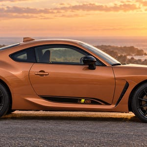 May include: A metallic orange two-door sports coupe with a sleek aerodynamic profile, black multi-spoke alloy wheels, and a black side rocker panel decal featuring the GR 86 logo. The vehicle is captured from a side profile view at sunset.