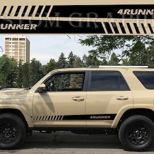 May include: Tan SUV with black accents and wheels. The side features a black stripe with the text "4Runner." Above the vehicle is a graphic with the text "4Runner" and diagonal black stripes. The background includes trees and a building.