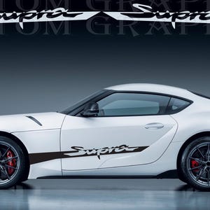 Puede incluir: Un coche deportivo blanco con ruedas negras y pinzas de freno rojas. El coche tiene una calcomanía "Supra" en blanco y negro en el lateral. La imagen también muestra una calcomanía "Supra" en blanco y negro encima del coche.