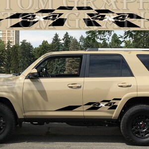 Op de afbeelding: Tan SUV met zwarte accenten en 4x4-stickers. Het voertuig heeft zwarte velgen en banden. De stickers zijn zwart-wit geblokte vlagontwerpen met de tekst "4x4". De achtergrond omvat bomen en een gebouw.