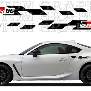 Puede incluir: Coche deportivo blanco con ruedas negras y gráficos laterales. Los gráficos incluyen formas de diamante negras y rojas y el texto "GR 86" en un cuadro blanco. El coche tiene un diseño elegante y moderno.