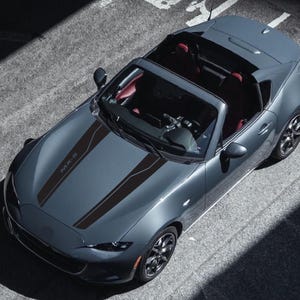 Puede incluir: Un Mazda MX-5 Miata convertible gris con rayas de carreras negras en el capó. El coche está aparcado en una superficie de asfalto gris.