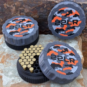 May include: Three circular storage containers with textured gray sides and lids featuring an orange, gray, and black camouflage pattern. Each lid is embossed with the text 22LR. One container is open, revealing a black honeycomb interior holding brass-colored 22LR cartridges.