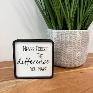 May include: A square sign with a black frame and a white face featuring the black text "NEVER FORGET THE difference YOU MAKE" in a combination of block and script fonts. The sign is positioned in front of a textured gray planter.