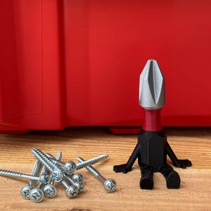 May include: A whimsical, black and silver screwdriver bit figure with a red neck, sitting on a wooden surface. A pile of silver screws is to the left. A red storage container is in the background.