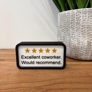 May include: A rectangular black-framed sign with a white background featuring five gold stars and the black text "Excellent coworker. Would recommend." The sign has a textured black border and is displayed in a horizontal orientation.