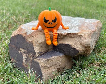May include: A bright orange pumpkin figurine with a green stem, featuring a black printed face with sharp, angry eyes and a jagged, stitched-style mouth. The pumpkin has articulated, segmented orange arms and legs extending from its body.