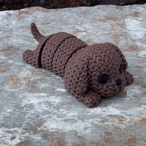May include: A brown, segmented dog figurine with a textured surface. The dog is lying down with its tail curved upwards. The eyes and nose are black. The figurine is resting on a gray and brown stone surface.