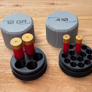 May include: Two gray cylindrical containers with the text "12 GA" and ".410" on top, next to black holders with red and gold ammunition. The holders have multiple holes to hold the ammunition. The items are on a wooden surface.