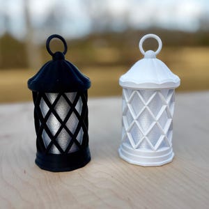 May include: Two decorative lanterns, one black and one white, with a diamond-shaped lattice design. The lanterns have a small handle at the top and are made of a smooth, shiny material.