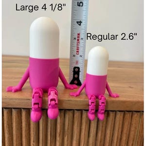 以下が含まれることがあります： 2つの気まぐれなピル型の置物。大きい方は10.5cm、小さい方は6.6cmです。上部は白、中央はピンク、脚は関節付きのピンク色です。「Large 4 1/8」と「Regular 2.6」の文字が見えます。