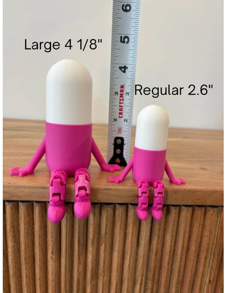 以下が含まれることがあります： 2つの気まぐれなピル型の置物。大きい方は10.5cm、小さい方は6.6cmです。上部は白、中央はピンク、脚は関節付きのピンク色です。「Large 4 1/8」と「Regular 2.6」の文字が見えます。