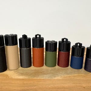May include: A collection of seven cylindrical containers with textured bodies in various colors, including black, tan, dark grey, orange, green, red, blue, and purple. Each has a black top with a unique design.