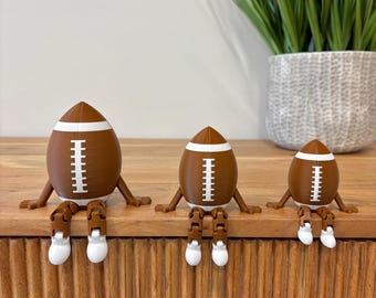 May include: Three brown and white American football-shaped figures of varying sizes, each featuring articulated brown arms and legs with white feet. The figures are positioned in a row, sitting upright with their legs dangling over the edge of a surface.